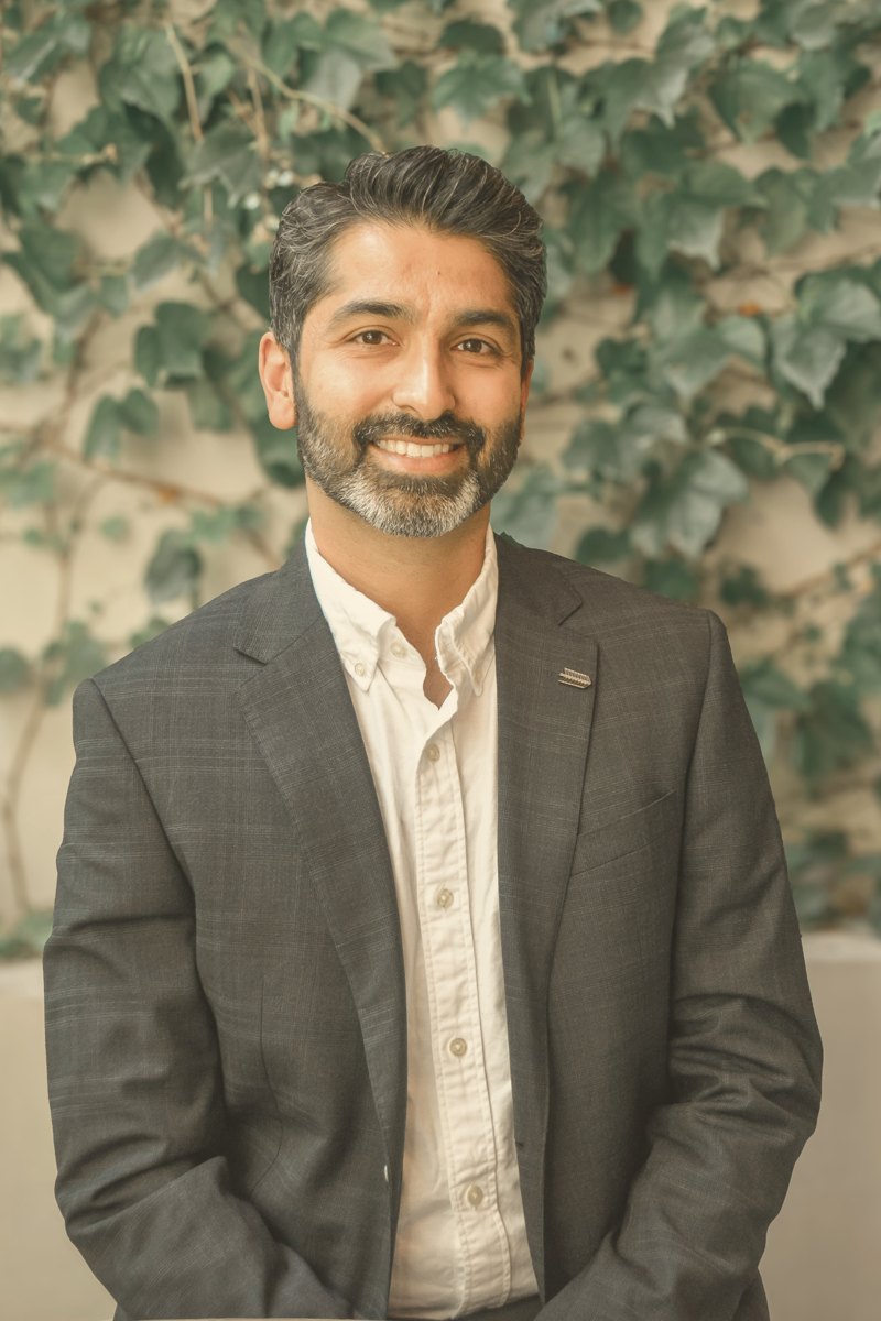 Dr Bharat Visa smiling, wearing a charcoal blazer and white shirt in front of an ivy-covered wall